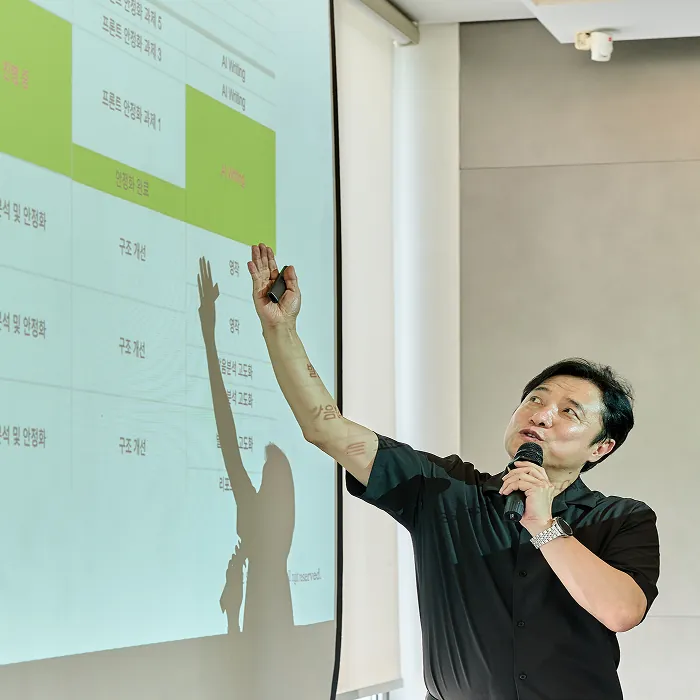 Han Sunghee holding a microphone and pointing to a table on screen during an Asan Sanghoe lecture